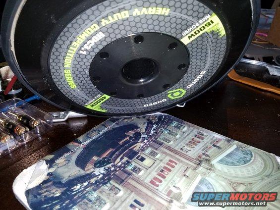 Car audio subwoofer with "HEAVY DUTY COMPETITION SERIES MDC3.1" text, next to a book showing the Library of Congress
