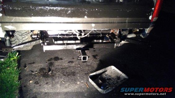 Rear of a dark-colored vehicle with a trailer hitch installed. A black pan with some debris sits on the asphalt
