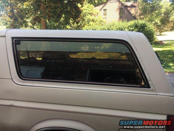 Side view of a 1988 Ford Bronco. The rear window reflects trees and a house