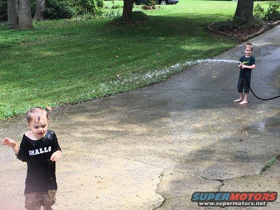 Young boy sprays water from a hose at another boy standing in a puddle