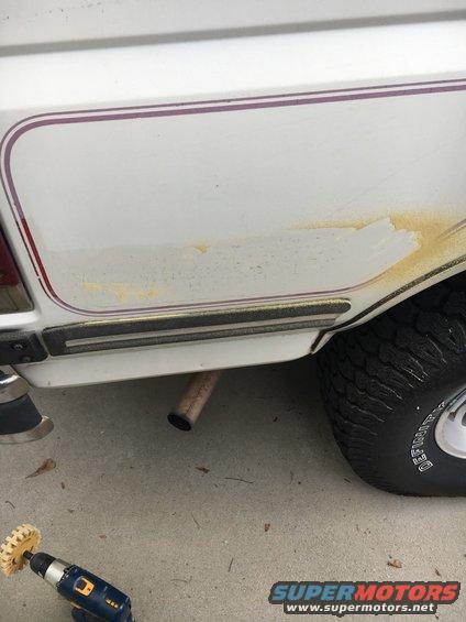 White SUV rear quarter panel with purple stripe, showing rust spots and an exhaust pipe. A drill with a brush attachment is on the ground