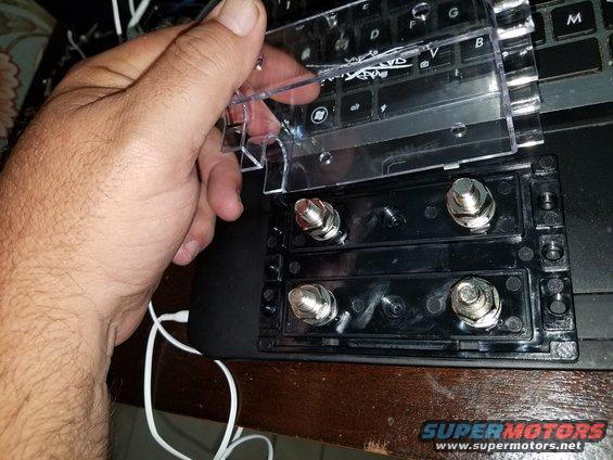 Hand holding clear plastic cover over black car stereo power distribution block with four silver bolts