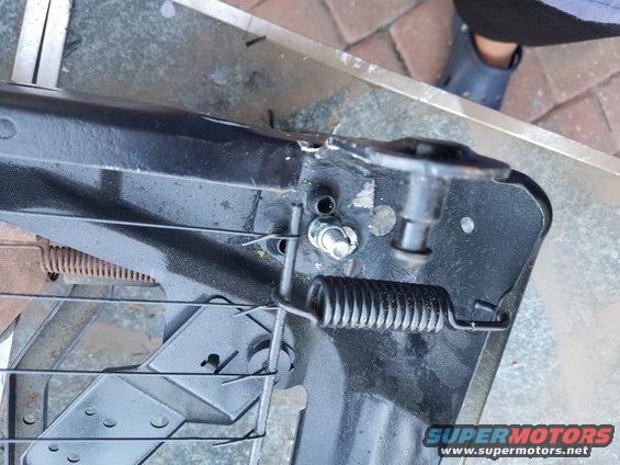 Close-up of a black metal seat frame with springs and bolts. A person's blue shoe is visible in the background