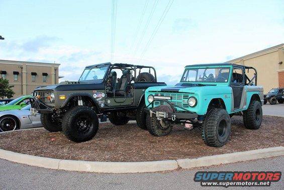 Two Ford Broncos, one olive green and one teal, parked side-by-side on a dirt patch with a building in the background