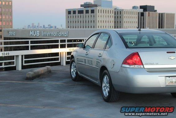 Silver sedan parked on a rooftop, rear quarter view. Building and city skyline in background