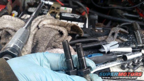 Two bolts held by a gloved hand, with engine components and tools in the background