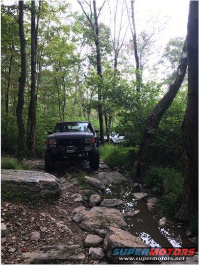 Dark-colored Ford Bronco driving on a rocky, wooded trail with a stream