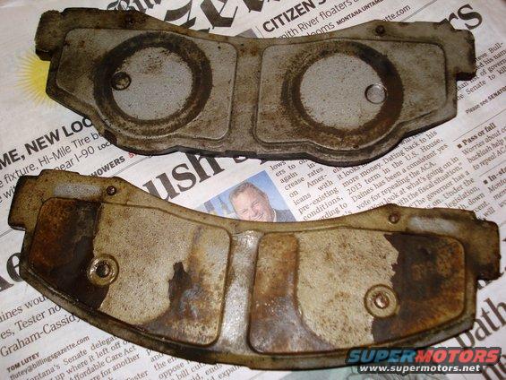 Two used, dirty brake pads with circular indentations and metal backing plates, resting on newspaper