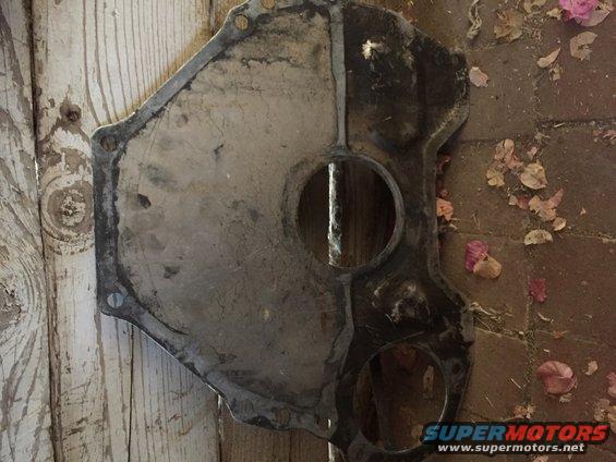 metal engine timing cover with circular cutouts, lying on wooden planks and brick patio with leaves