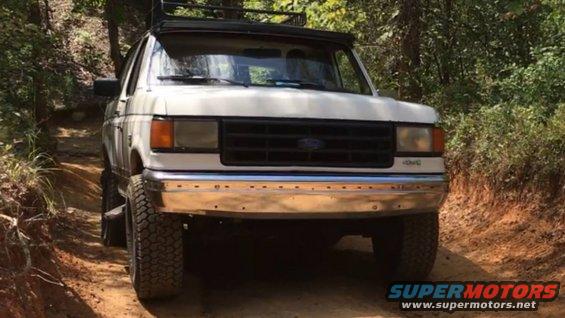 White Ford Bronco on a dirt road with trees on either side. It has a roof rack