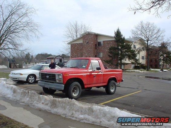 Red Ford Bronco parked in a lot, 3/4 front view. White car to the left