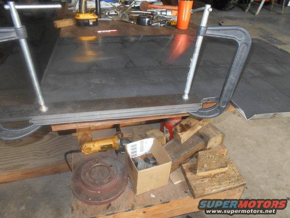 Metal sheet clamped to wooden surface with angle grinder and brake rotor nearby