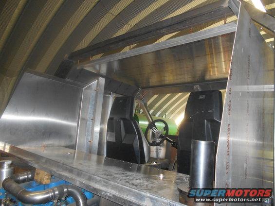 Interior view of a custom vehicle build, showing unfinished metalwork, two bucket seats, and a steering wheel