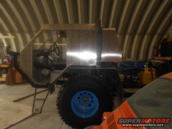 Custom vehicle build in a garage, showing unfinished metal body panels and a large tire with a blue rim
