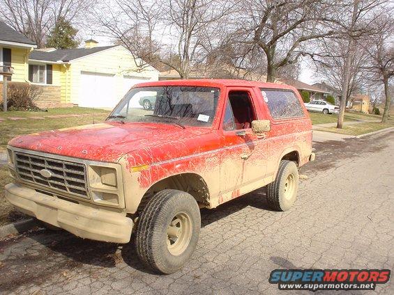 Mud-covered red 1980 Ford Bronco parked on a cracked asphalt street
