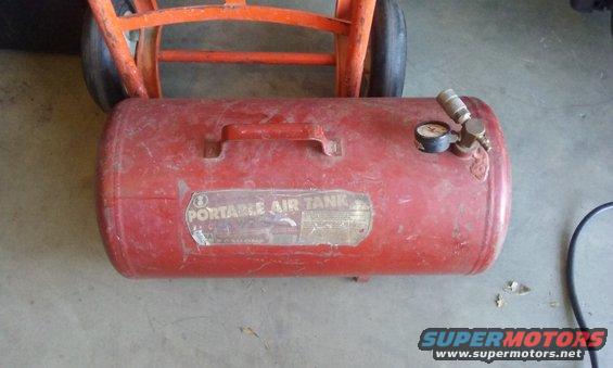 Red portable air tank with a gauge and handle, resting on an orange hand truck