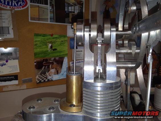 Close-up of a model engine's crankshaft and piston mechanism. Photos and labels are pinned to a corkboard in the background