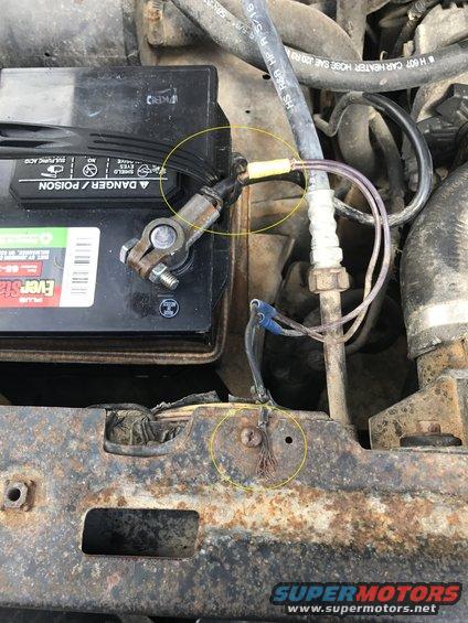 Car battery with corroded terminals and frayed wires, surrounded by engine components and rusty metal