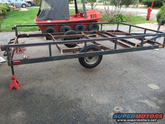 Utility trailer frame with wheels, supported by red jack stands. Red amphibious vehicle in background