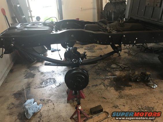 Truck frame with leaf springs and rear axle on jack stands. Black painted metal, some fluid stains on concrete floor