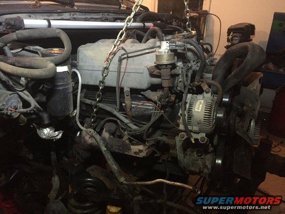 Engine assembly suspended by chains, showing alternator, hoses, and wiring. Some parts are dirty and worn