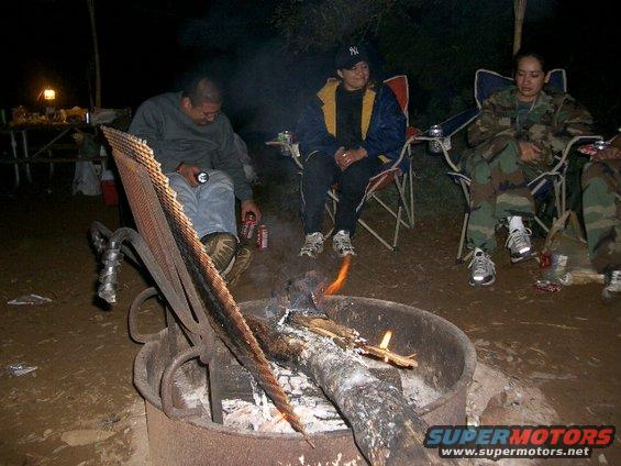 People sit around a campfire at night, with a picnic table and lantern visible in the background