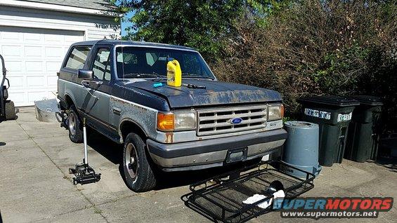 1987 Ford Bronco, dark blue and gray, with a yellow oil jug on the hood, parked in a driveway