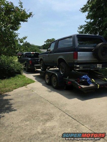 1987 Ford Bronco on a trailer, being towed by a black pickup truck