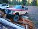 1987 Ford Bronco II - Two off-road vehicles navigate a challenging path through fallen logs in a wooded area.