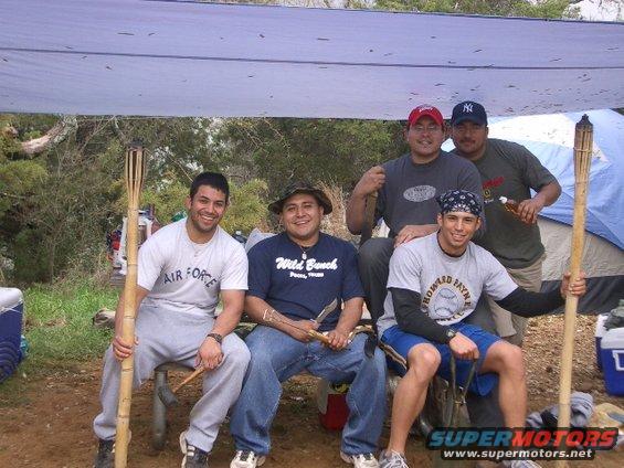 Five men pose for a photo under a tarp at a campsite. One man wears an 'Air Force' shirt, another a 'Wild Bunch' shirt