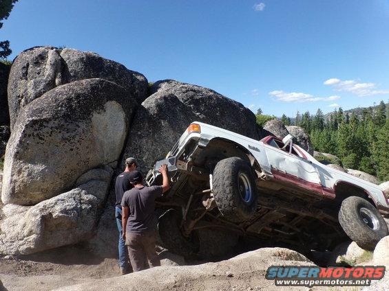 White and red off-road vehicle tilted on rocks with people nearby