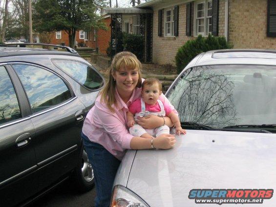 Woman holding a baby on the hood of a silver car, with a dark SUV parked next to it