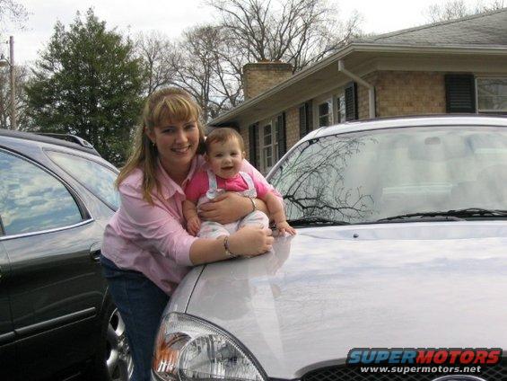 Silver 2004 Ford Taurus with a woman and baby on its hood, parked next to a dark SUV