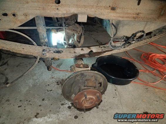 Underside of a vehicle showing a leaf spring, rear axle, and brake drum assembly. A drain pan with fluid and orange extension cords are nearby