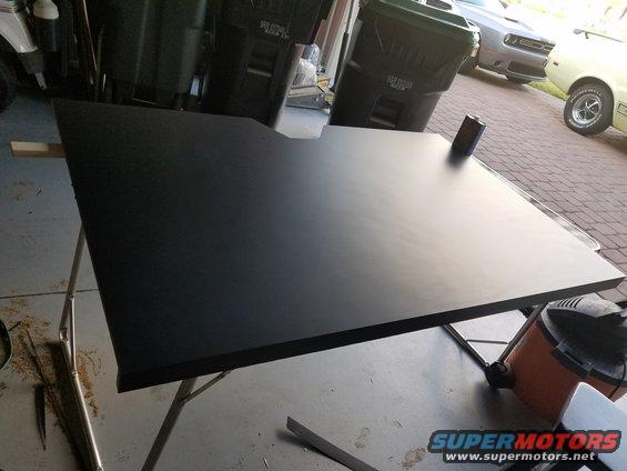 Black dashboard panel on sawhorses in a garage, with trash cans and cars in the background