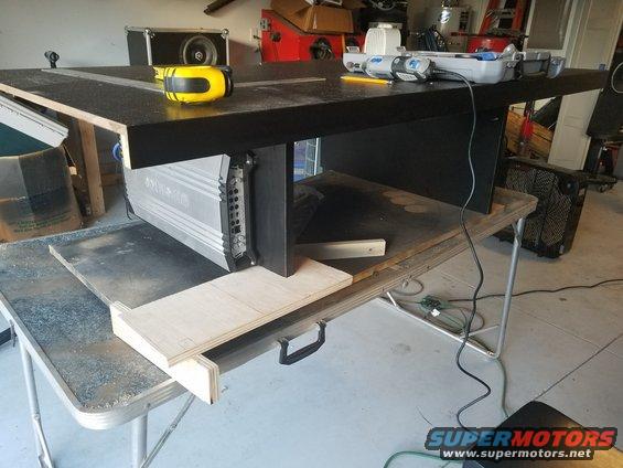 Silver car amplifier mounted under a black table, with a yellow tape measure on top