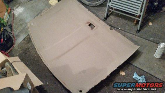 Car headliner removed and lying on a garage floor. It has a rectangular cutout for a light or speaker