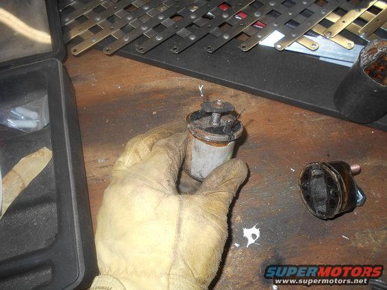 Gloved hand holding a dirty automotive starter solenoid; another part is on the table