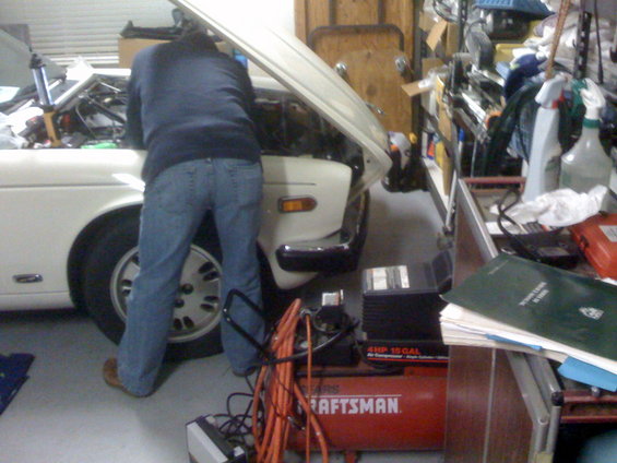Person in jeans working on car engine in garage with air compressor and tools nearby