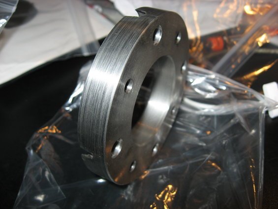 Metal flange with six bolt holes, brushed finish, sitting in plastic packaging
