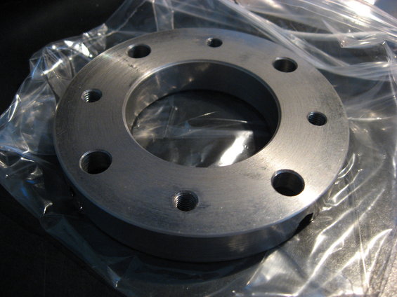 Metal flange with six bolt holes, some threaded, in plastic packaging
