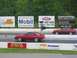 1986 Ford Mustang - Two red 1986 Ford Mustangs compete on a drag strip, with various sponsor advertisements visible.