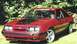 1986 Ford Mustang - A red coupe with a black hood accent and 'MOTORSPORT' banner is parked on grass with a wooded background.