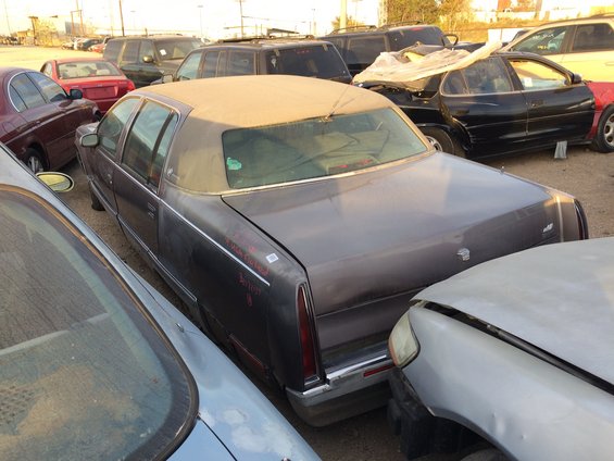 Dark sedan with a vinyl top parked in a junkyard, surrounded by other vehicles. Red writing on the trunk