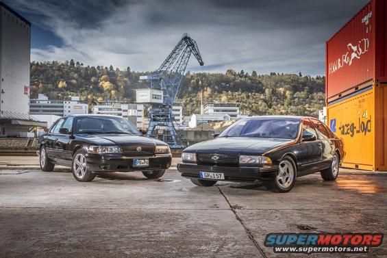 Black Mercury Marauder and a black Chevrolet sedan parked side-by-side in an industrial area with cranes and shipping containers