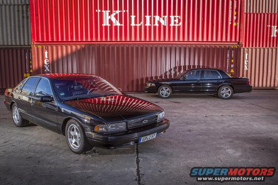 Two black sedans parked in front of red shipping containers. One is a Chevrolet Caprice, the other is a Ford Crown Victoria
