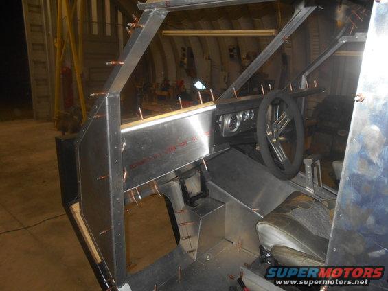 Interior of a custom-built vehicle under construction, showing unfinished metal panels, dashboard, steering wheel, and seat