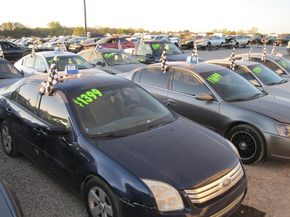 Several cars parked in a lot, some with checkered flags and price tags. One dark blue car in foreground has $1399 on windshield