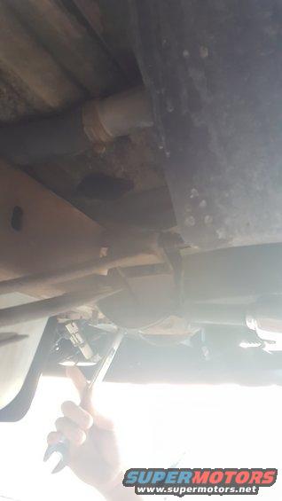 Underbody of a vehicle with a hand using a wrench on a bolt. A Supermotors.net logo is visible