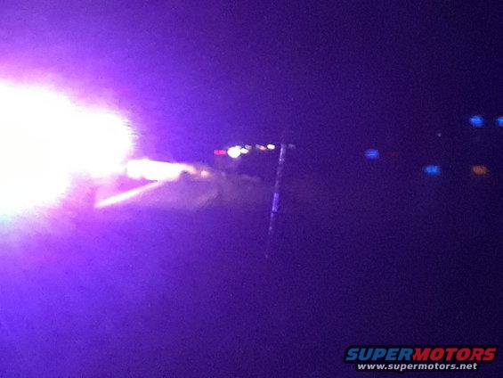 Night scene with bright purple and white lights illuminating a road, possibly after an accident. Distant lights are visible